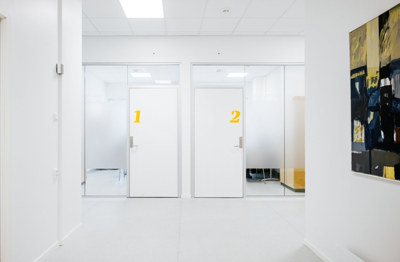 Interiør fra helsestasjon for ungdom. Hvite veggflater med et maleri, glassvegger og to hvite dører med de gule tallene 