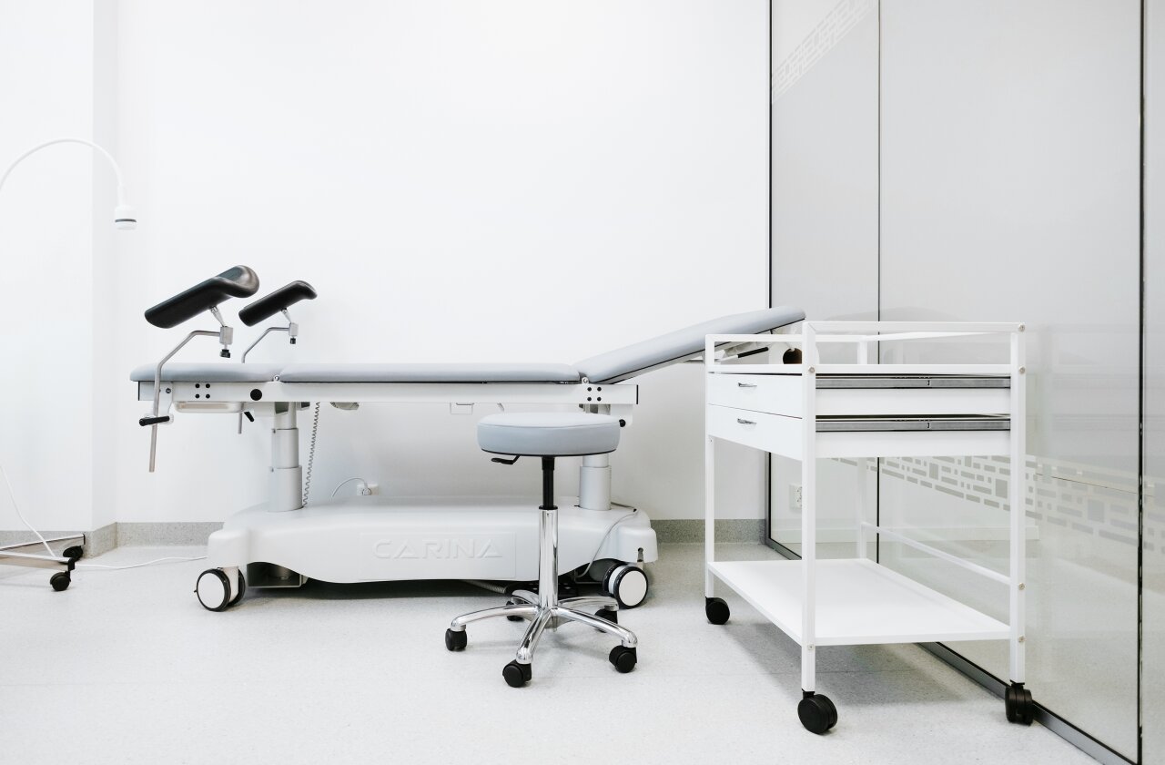 Interiør fra et legekontor med pasientseng en krakk på jul til legen og et trillebord til utstyr.
