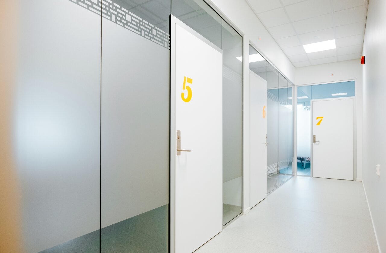 Interiør fra helsestasjon for ungdom. Hvite veggflater, glassvegger og tre hvite dører med de gule tallene 