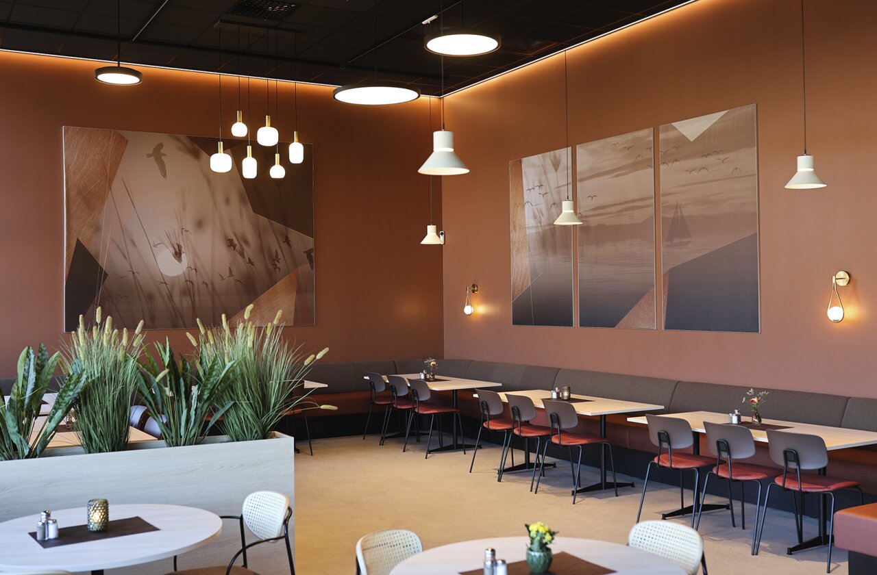 Kafélokale med flere sittegrupper, grønne planter og lun belysning. På de beige veggene henger store tekstilbilder med naturfoto i beige toner.